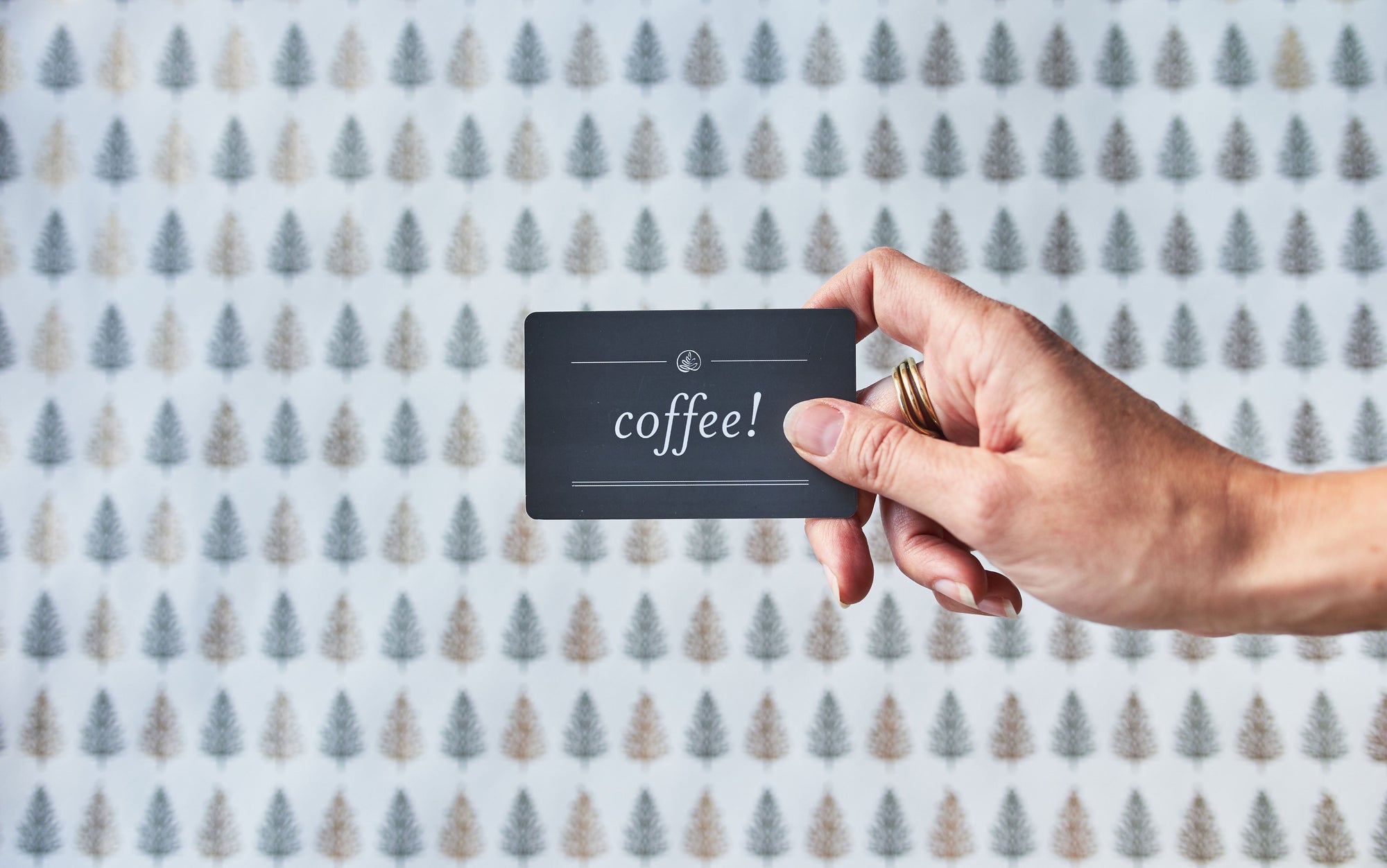 a hand holding a gift card over christmas tree themed wrapping paper. the gift card is grey with white text which reads coffee