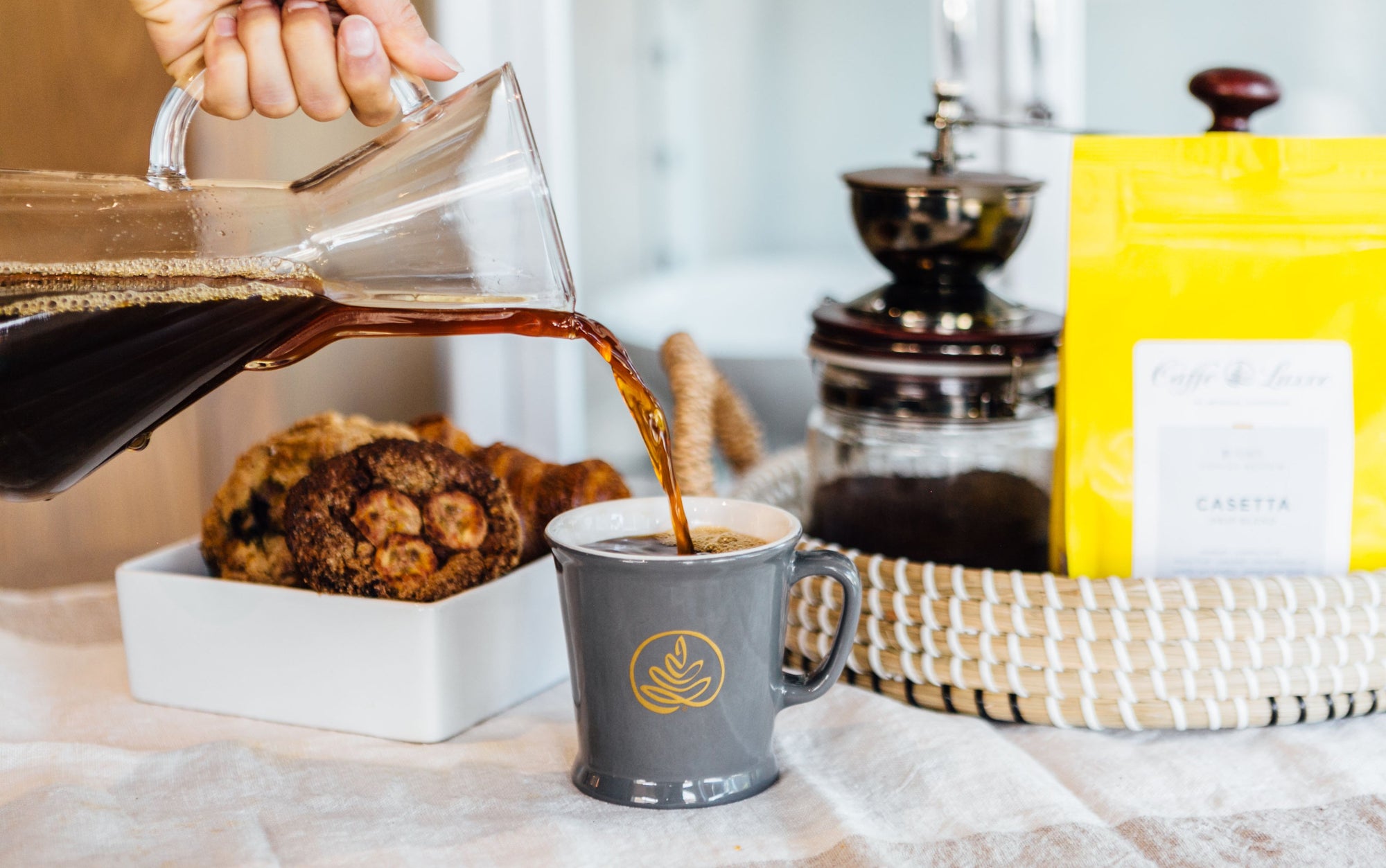 coffee being poured from a small 1 - 3 cup chemex into a grey mug, which bears a caffe luxxe logo in gold. in the background are pastries, a bag of coffee, and a manually operated grinder.